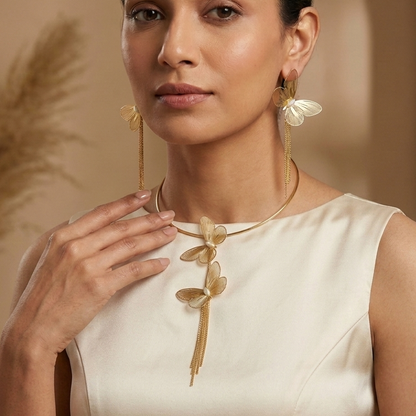 A woman wearing a gold necklace with a leaf design and matching long drop earrings, posed against a warm beige background with a pampas grass prop.